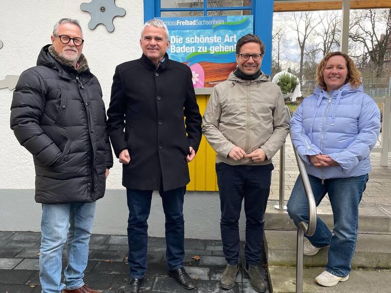 Vier Personen stehen lächelnd vor einem Gebäude mit einem bunten Fenster und einem Schild im Hintergrund.