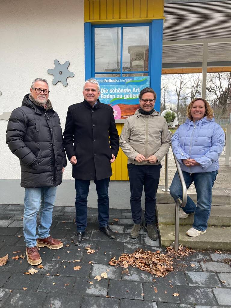 Vier Personen stehen lächelnd vor einem Gebäude mit einem bunten Fenster und einem Schild im Hintergrund.
