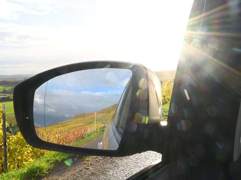 Der Seitenspiegel eines Autos zeigt eine ländliche Landschaft mit grünen Feldern und Hügeln, während die Sonne blendend von oben hereinstrahlt.