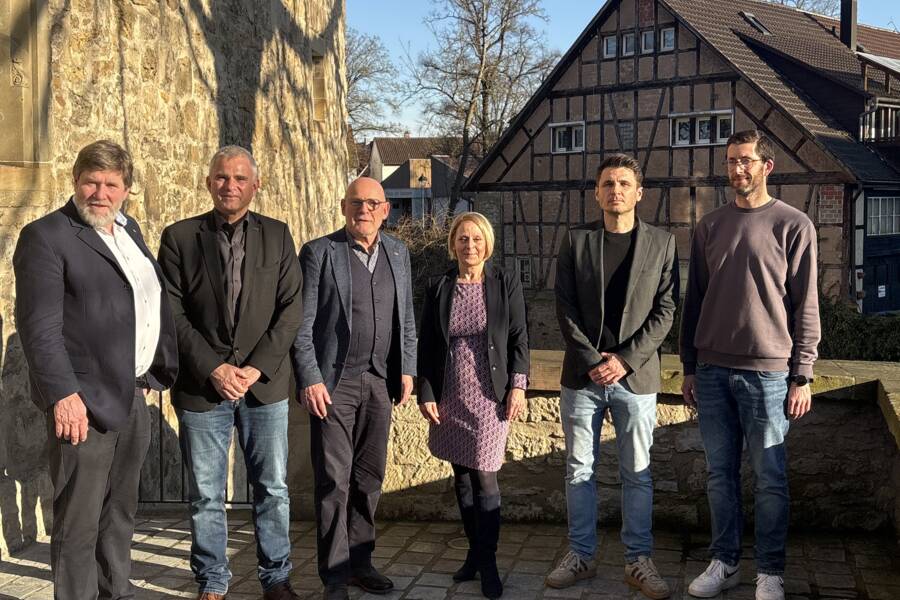 Eine Gruppe von sechs Personen steht nebeneinander auf einem gepflasterten Weg vor einem Gebäude mit Fachwerk, bei sonnigem Wetter.