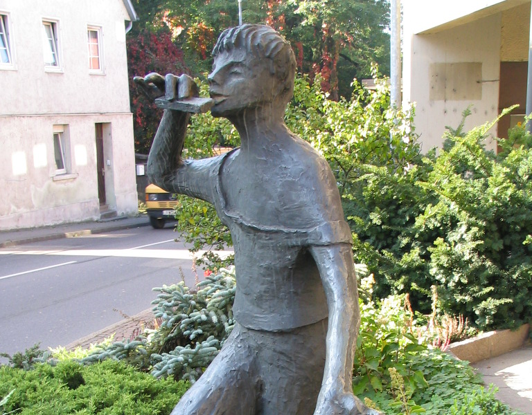 Eine Bronzestatue zeigt einen Mann mit einer Flöte und einer Ente, der in einer grünen Gartenlandschaft steht.