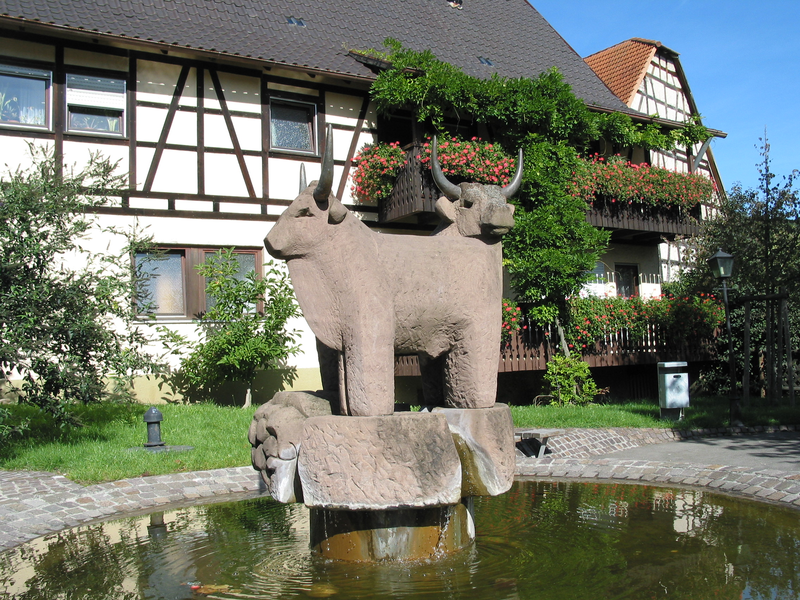 Eine große Steinskulptur zweier Ochsen steht in einem kleinen Teich vor einem Fachwerkhaus mit bepflanztem Balkon.