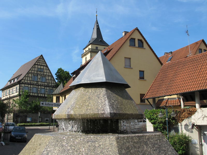 Ein modern gestalteter, mehrstufiger Brunnen mit fließendem Wasser steht vor traditionellen Fachwerk- und Wohnhäusern unter einem klaren blauen Himmel.