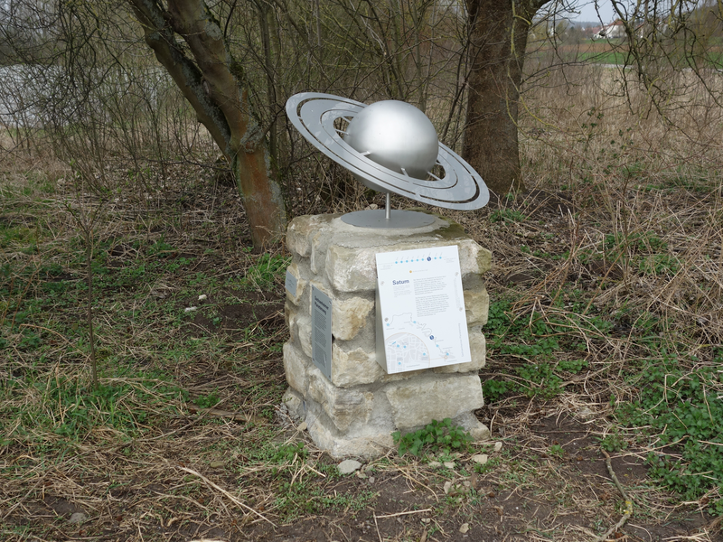 Eine kleine Metallskulptur des Planeten Saturn mit Ringen ist auf einem steinernen Sockel in einer bewachsenen Landschaft angebracht.