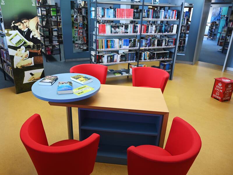 Eine Bibliothek mit Bücherregalen im Hintergrund, in der Mitte stehen ein runder Tisch mit Büchern und vier rote Stühle.