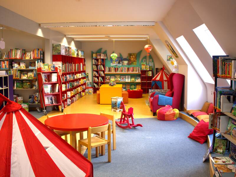 Ein bunter, gemütlicher Kinderbereich in einer Bibliothek mit Bücheregale, einem runden Tisch mit Stühlen, Kissen, einem Spielzelt und Spielzeug.