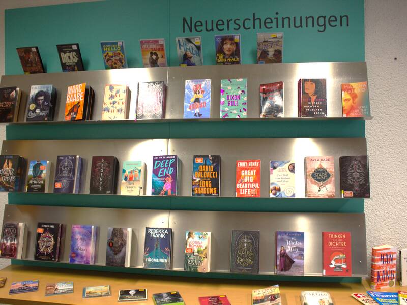 Ein Regal voller farbenfroher Buchcover unter dem Schild "Neuerscheinungen" in einer Bücherei.