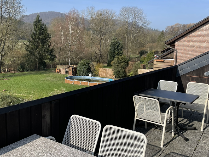 Eine Terrasse mit mehreren Tischen und Stühlen bietet Blick auf einen grünen Garten und Bäume vor einem klaren, blauen Himmel.
