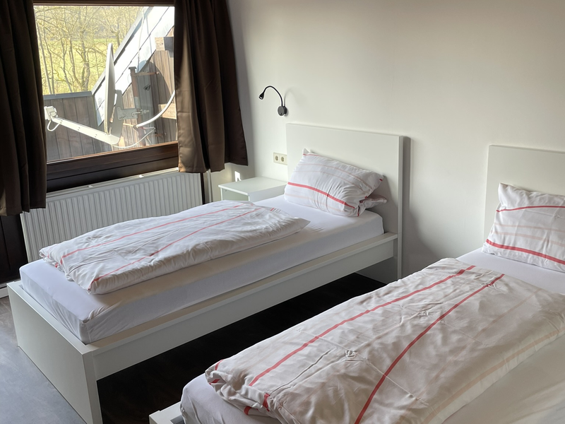 In einem Zimmer mit Holzbalkendecke stehen zwei Einzelbetten mit weißer Bettwäsche und roten Streifen nebeneinander, vor einem großen Fenster mit braunen Vorhängen.