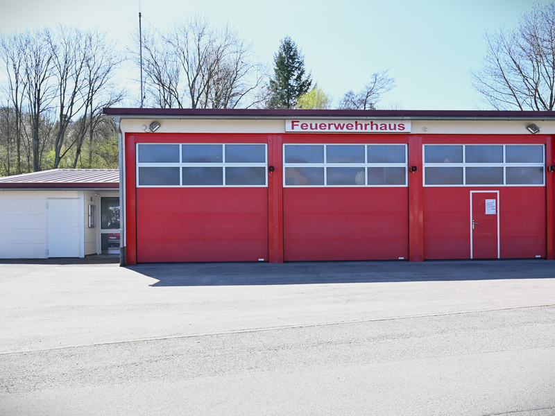 Das Bild zeigt ein rotes Feuerwehrhaus mit drei großen Toren und einem Schild darüber, auf dem "Feuerwehrhaus" steht, umgeben von Bäumen im Hintergrund.