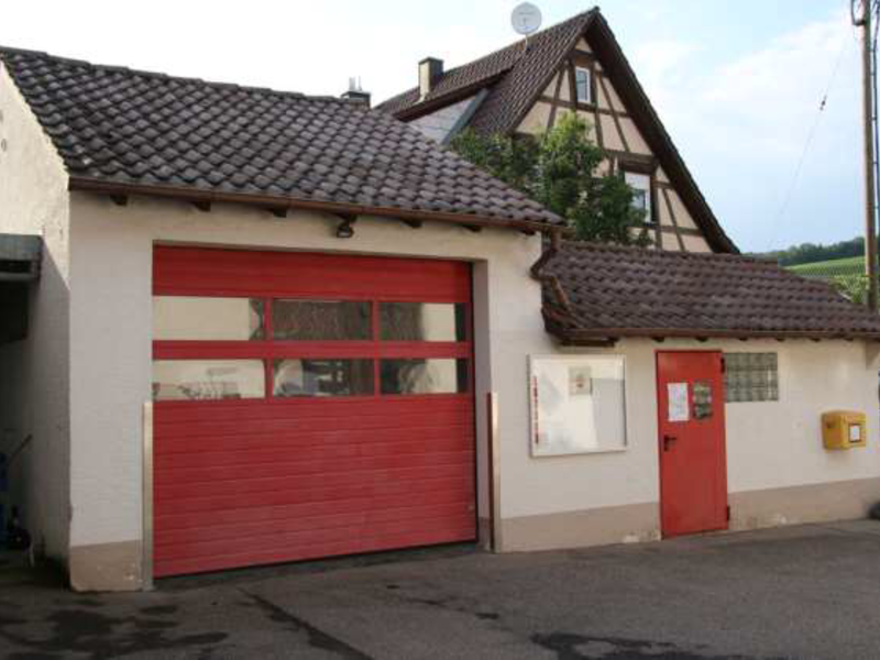 Ein kleines Gebäude mit einem roten Garagentor und einer roten Tür, daneben steht ein größeres Fachwerkhaus.
