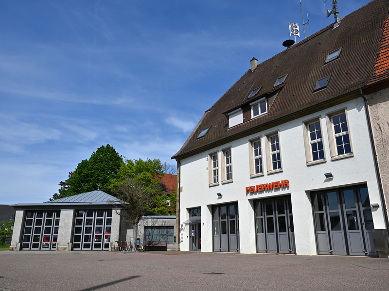 Ein Feuerwehrgebäude mit mehreren Toren und einem Schild mit der Aufschrift "FEUERWEHR" an einer weißen Fassade unter einem blauen Himmel.
