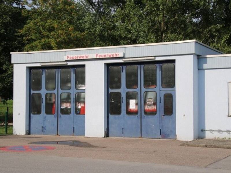 Ein kleines, hellblaues Feuerwehrgebäude mit zwei großen blauen Toren und einem Schild mit der Aufschrift "Feuerwehr" darauf.