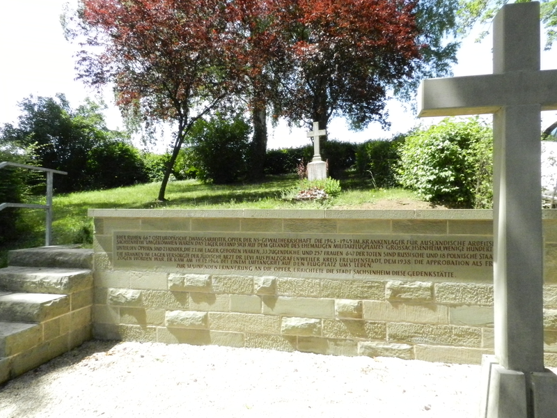 Eine Gedenkstätte mit einem großen Kreuz, einer Inschrift auf einer Steinmauer und Bäumen im Hintergrund.