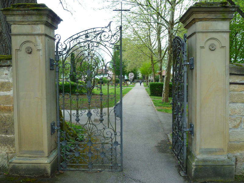 Ein kunstvoll verziertes, schmiedeeisernes Tor öffnet sich zu einem von Bäumen gesäumten Weg in einem FriedhofsgGelände.
