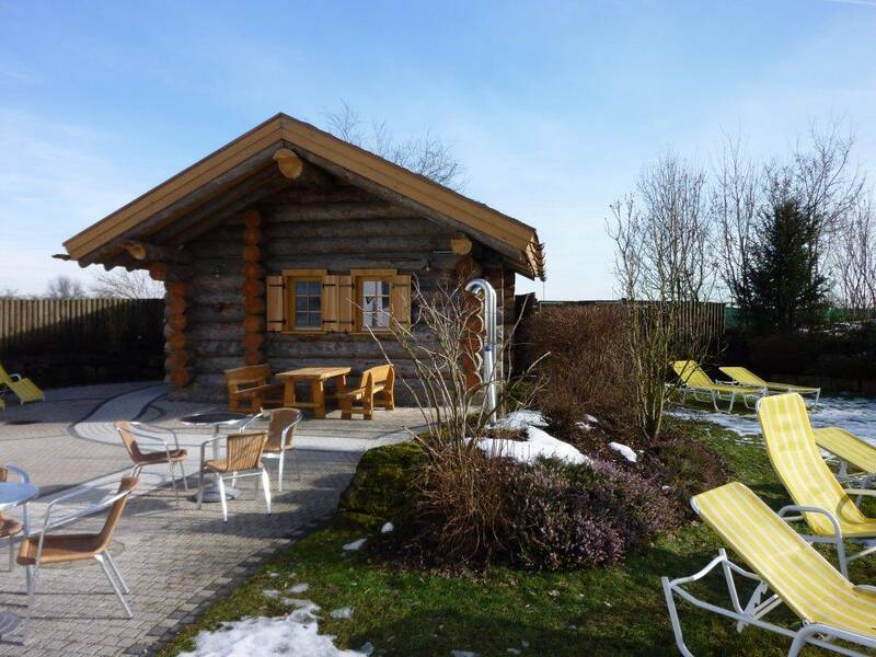 Eine kleine Blockhütte mit schneefreiem Gartenbereich, umgeben von Liegestühlen und Tischen mit Stühlen auf einer gepflasterten Fläche.