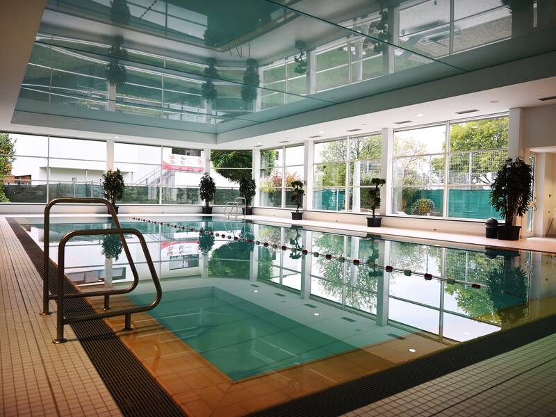 Ein Hallenbad mit hellen Fenstern, Pflanzen am Rand und einem klaren, ruhigen Wasserbecken.