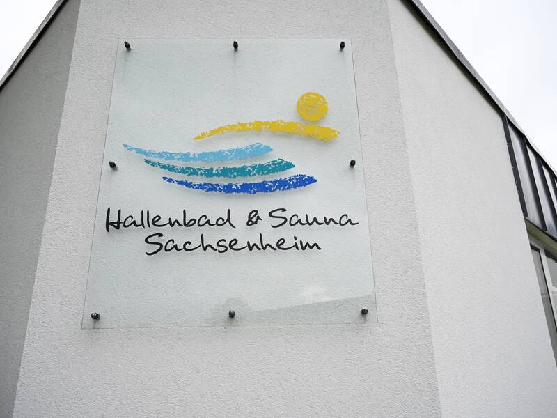 Ein Schild mit der Aufschrift "Hallenbad & Sauna Sachsenheim" ist an einer weißen Gebäudewand angebracht, verziert mit stilisierten blauen und gelben Wellen und einer Sonne.