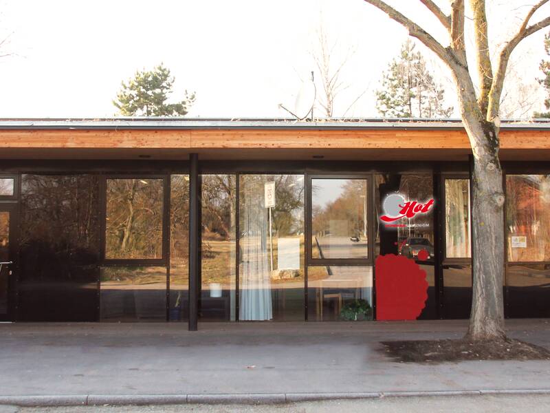 Das Bild zeigt die Außenansicht eines Gebäudes mit modernen Holz- und Glaselementen, vor dem ein Baum steht.