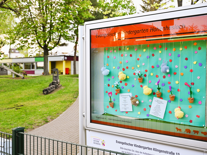 Ein Schaukasten des Evangelischen Kindergartens Hohenhaslach zeigt eine frühlingshafte Dekoration mit bunten Blumen und Tieren vor einer grünen Kulisse.