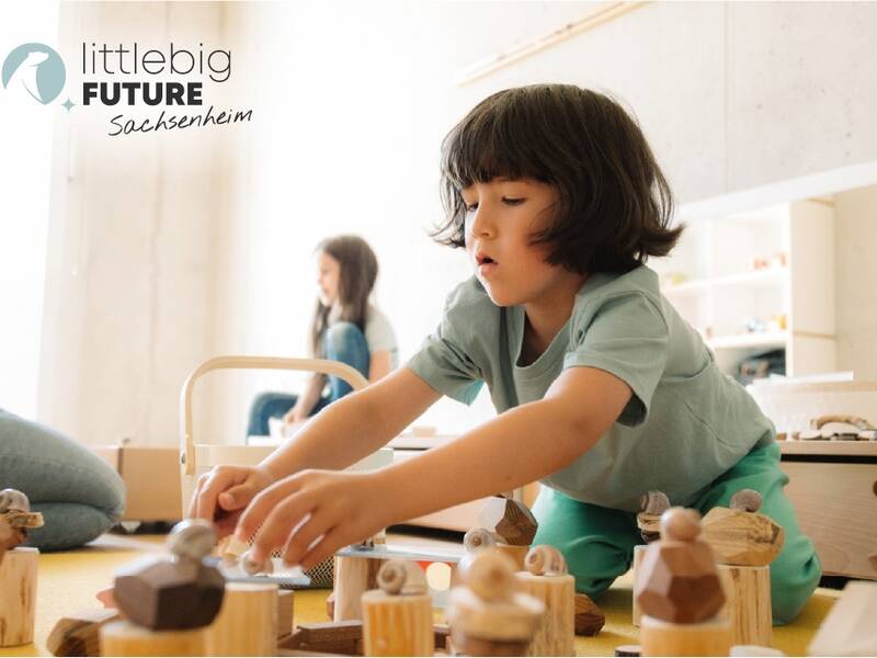 Ein Kind spielt konzentriert mit Holzspielzeug auf einem Boden, während weitere Menschen im Hintergrund zu sehen sind; oben links ist das Logo von "littlebig FUTURE Sachsenheim" zu sehen.