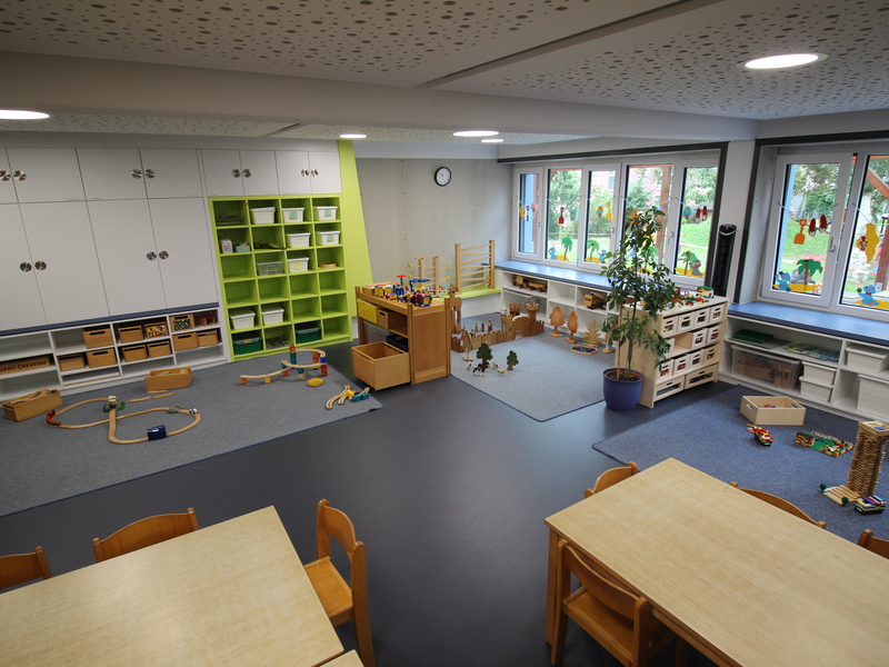 Ein heller Kindergartenraum mit Regalen voller Spielzeug, Bauklötzen und Malutensilien, sowie kindgerecht gestalteten Tischen und Stühlen.