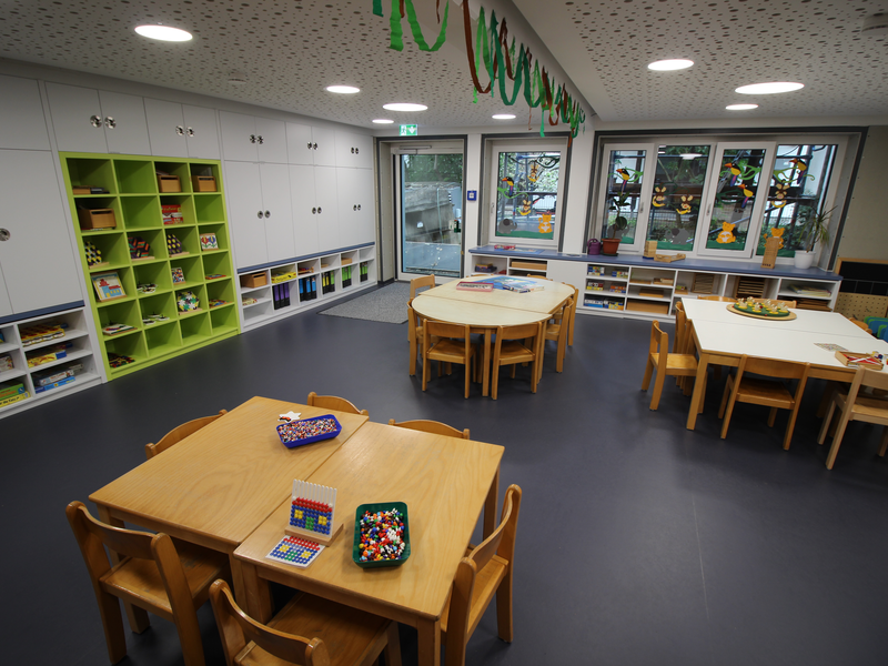 Ein heller, moderner Kindergartenraum mit mehreren Tischen und Stühlen, Regalen voller Spiele und Büchern sowie großen Fenstern, die zum Spielen und Lernen einladen.