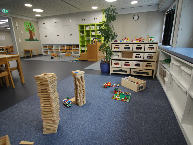 Ein heller Kindergartenraum mit Spielecken, Bauklötzen und Spielzeugregalen, in dem ein hoher Turm aus Holzbausteinen gebaut wurde.
