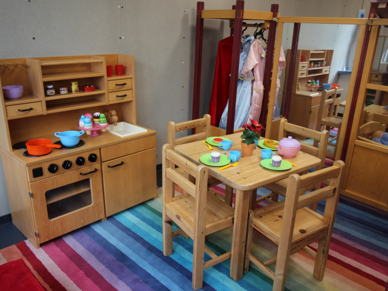 Ein Spielbereich mit einer hölzernen Kinderspielküche und einem Tisch mit vier Stühlen, der mit Spielgeschirr gedeckt ist, steht auf einem bunten gestreiften Teppich.
