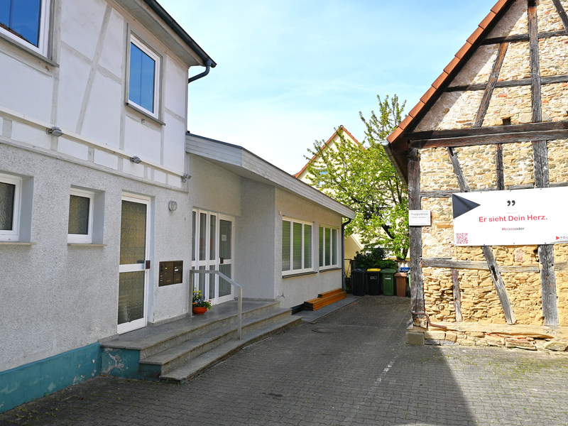 Zwei Gebäude, eines aus Beton und eines im Fachwerkstil, stehen nebeneinander, wobei das Fachwerkhaus ein Schild mit der Aufschrift "Er sieht Dein Herz" trägt.