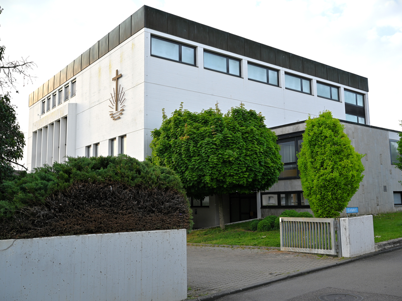 Ein großes, modernes Gebäude mit mehreren Fenstern und einer religiösen Symbolik an der Fassade, umgeben von gepflegtem Grün.