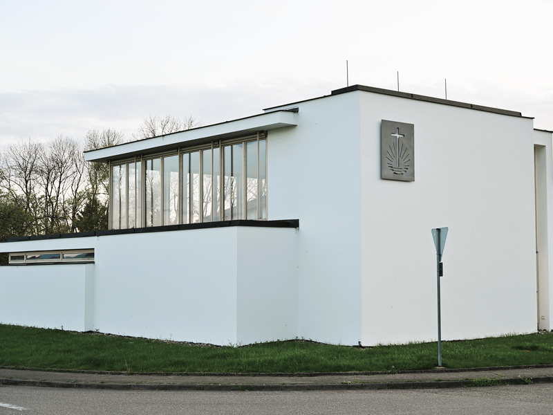 Das Bild zeigt ein modernes, schlichtes weißes Gebäude mit großen Glasfenstern und einem Kreuzemblem an der Fassade, umgeben von einer grünen Wiese.