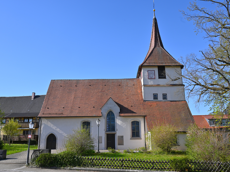Eine kleine Kirche mit rotem Ziegeldach und einem Kirchturm steht bei klarem Himmel in einer grünen Umgebung.