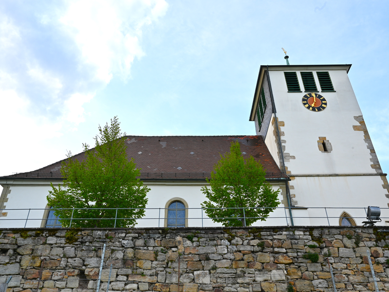 Die Aufnahme zeigt eine Kirche mit einem Turm, auf dem eine Uhr zu sehen ist, flankiert von Bäumen vor einer Steinmauer.
