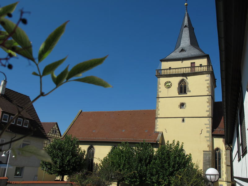 Das Bild zeigt einen gelben Kirchturm mit Ziffernblatt und spitzer Dachhaube, umgeben von weiteren Gebäuden und Bäumen unter einem klaren blauen Himmel.