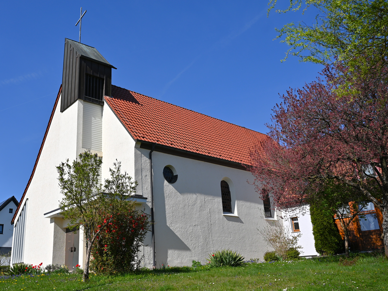 Eine kleine weiße Kirche mit rotem Ziegeldach steht vor einem klaren blauen Himmel, umgeben von blühenden Bäumen und Sträuchern.