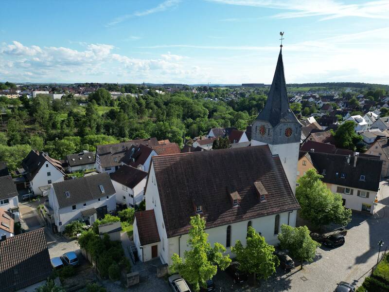 Luftaufnahme einer kleinen Stadt mit einer Kirche im Vordergrund, deren Turm mit einer Wetterfahne verziert ist, umgeben von Wohnhäusern und viel Grün.