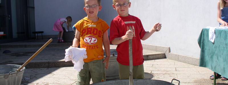 Zwei Jungen stehen vor einem großen Zuber mit Wasser und Schaumbürsten, während zwei weitere Kinder im Hintergrund spielen.