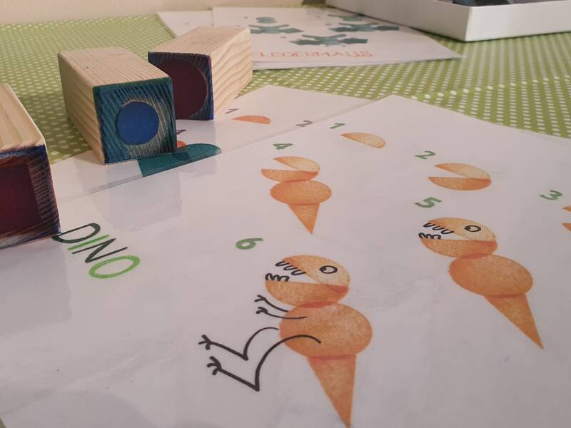 Auf einem Tisch liegen Holzblöcke und mehrere Papierblätter, die mit orangefarbenen Dinosaurierzeichnungen und Zahlen von 1 bis 6 bedruckt sind.