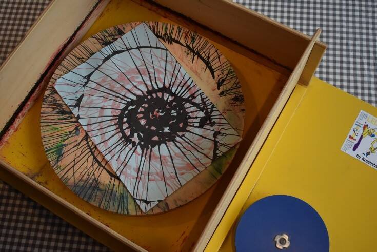 In einer Holzkiste liegt ein quadratisches Blatt Papier mit einem spiralförmigen, bunten Muster, das durch rotierende Farbspritzer entstanden ist.