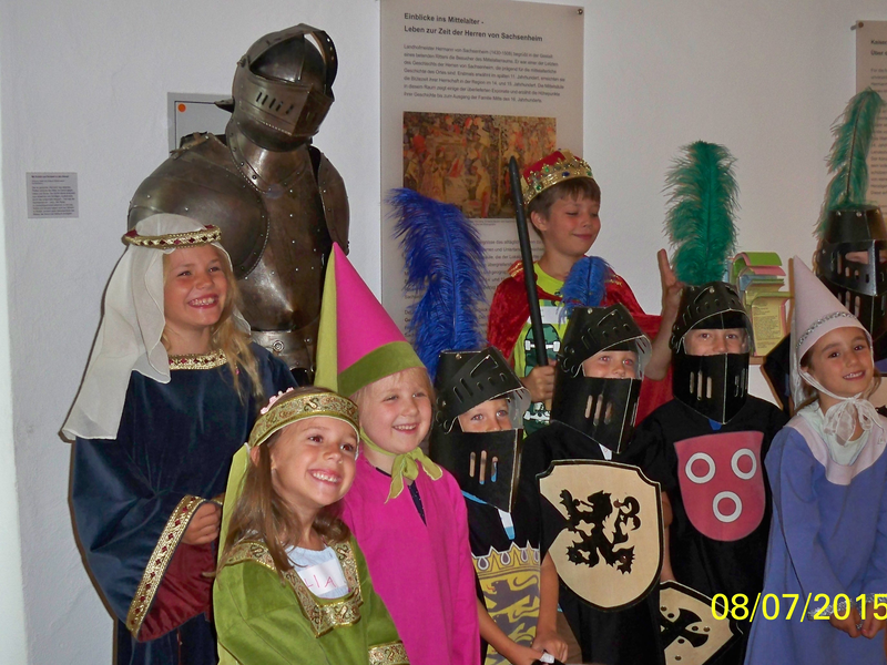 Eine Gruppe von Kindern ist fröhlich verkleidet als mittelalterliche Figuren, darunter Ritter und Prinzessinnen, und posiert neben einer Ritterrüstung.