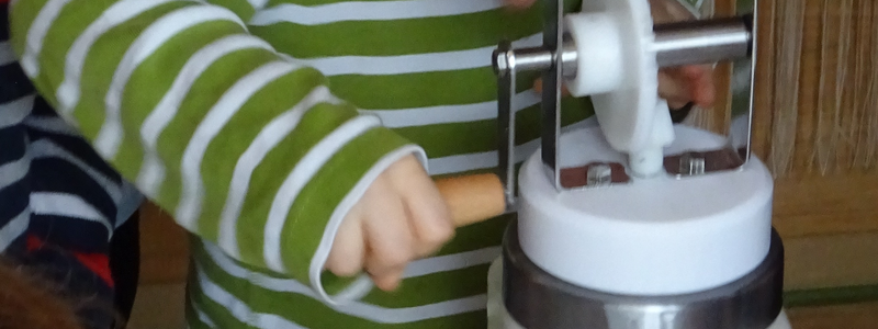 Ein Kind in einem gestreiften Pullover dreht konzentriert an einer Handkurbel, die mit einem Glasbehälter verbunden ist.