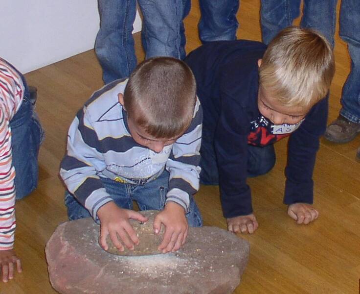 Zwei junge Kinder knien auf einem Holzboden und mahlen etwas mit einem runden Stein auf einem großen, flachen Stein, während andere Kindern im Hintergrund stehen.