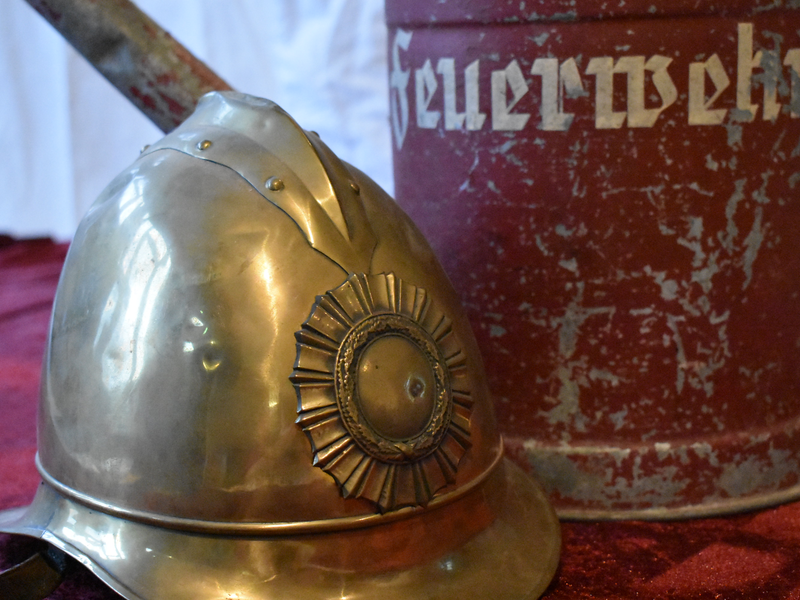 Ein alter Feuerwehrhelm aus Metall liegt neben einem rostigen, roten Behälter mit der Aufschrift "Feuerwehr".