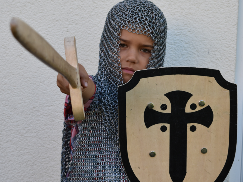 Ein Kind trägt ein Kettenhemd mit einem Helm und hält ein hölzernes Schwert und einen Schild mit einem Kreuzsymbol.
