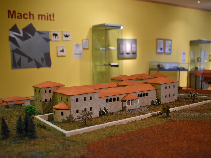Das Bild zeigt ein Architekturmodell einer antiken Villa in einem Ausstellungsraum mit gelben Wänden und der Aufschrift "Mach mit!".
