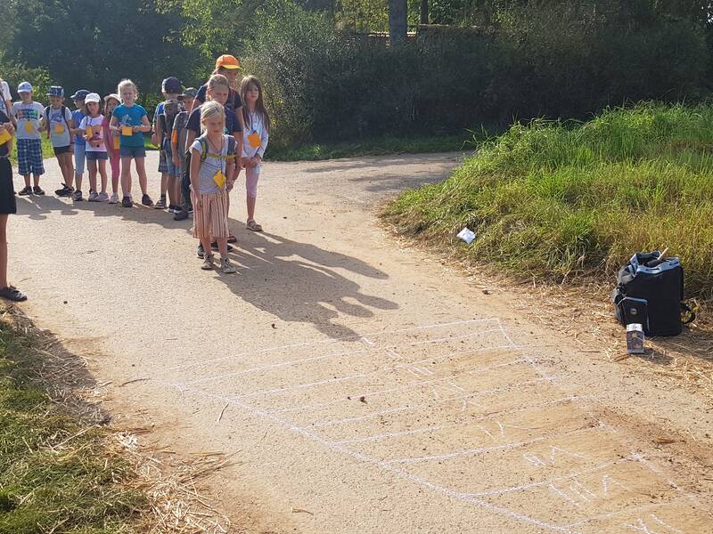 Eine Gruppe von Kindern steht auf einem Feldweg und schaut auf ein mit Kreide auf den Boden gemaltes Spielfeld, während einige der Kinder bunte Hüte und Schilder tragen.