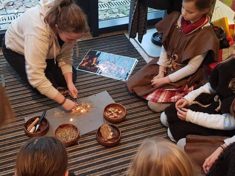 Eine Gruppe von Kindern sitzt auf einem Teppichboden um eine Person, die mithilfe von Stroh und Werkzeugen Feuer macht.