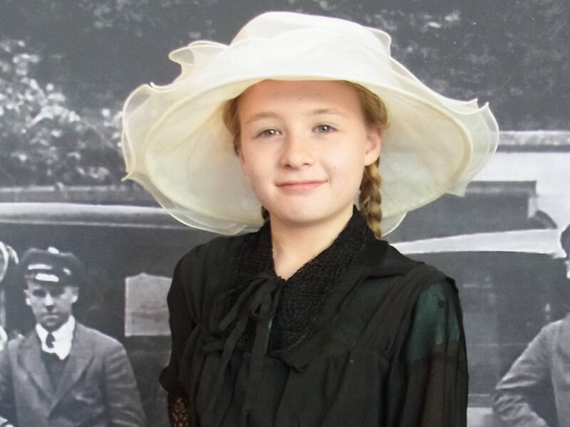 Ein junges Mädchen in einem schwarzen Kleid trägt einen weißen Hut und steht vor einem historischen, schwarz-weißen Foto mit Männern in alten Anzügen.
