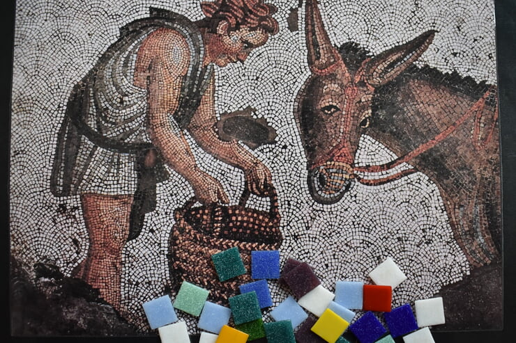 Ein Mosaik zeigt eine Person, die einem Esel einen Korb präsentiert, während bunte Mosaiksteinchen im Vordergrund liegen.